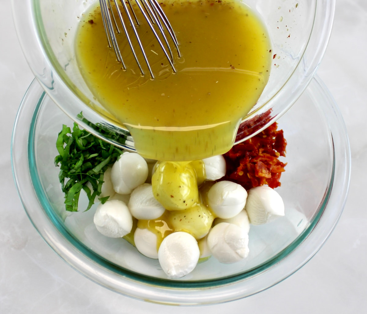 glass bowl with mozzarella balls, sun-dried tomatoes, chopped basil and parsley with marinade being poured over