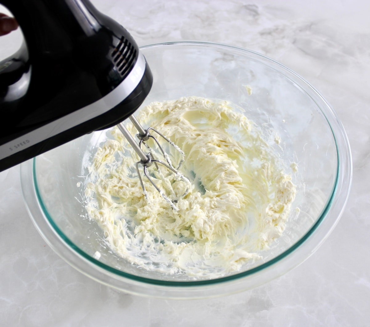 cream cheese in glass bowl being whipped with hand mixer