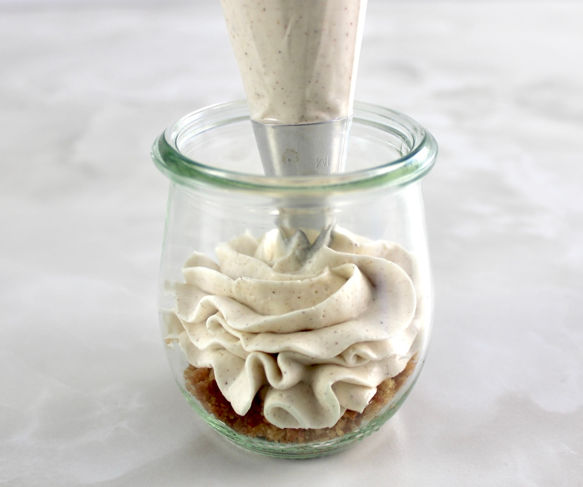 Keto Snickerdoodle Cheesecake fluff being piped into glass jar with cookie crumbs on bottom