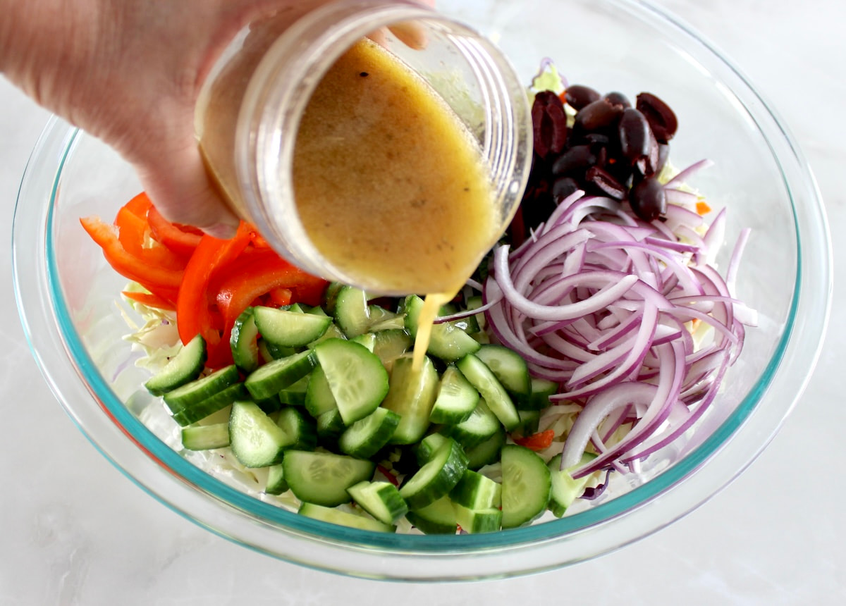 Greek salad dressing being poured over Greek Coleslaw in glass bowl