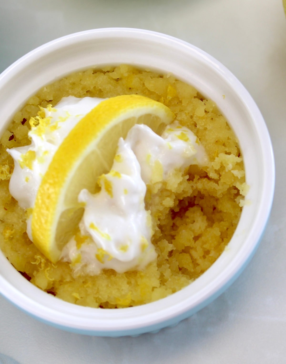 Keto Lemon Mug Cake in blue ramekin with whip cream, lemon slice and bite missing