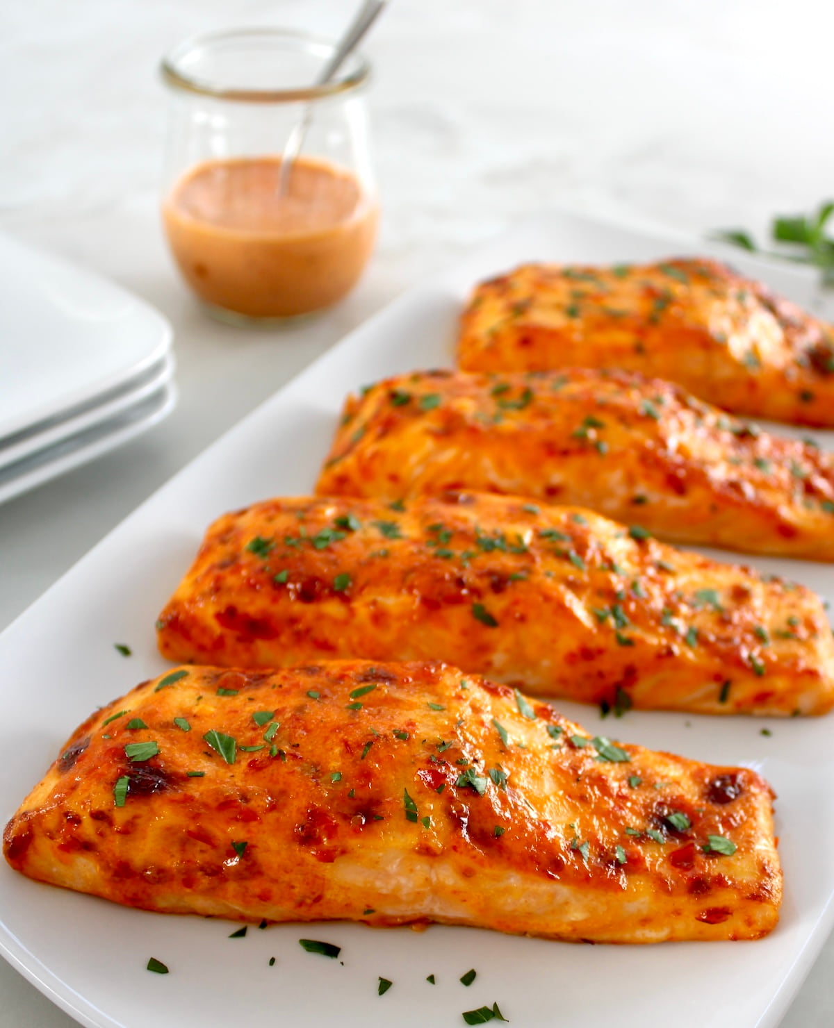 4 pieces of Bang Bang Salmon on white platter with chopped parsley on top and more sauce in jar in back