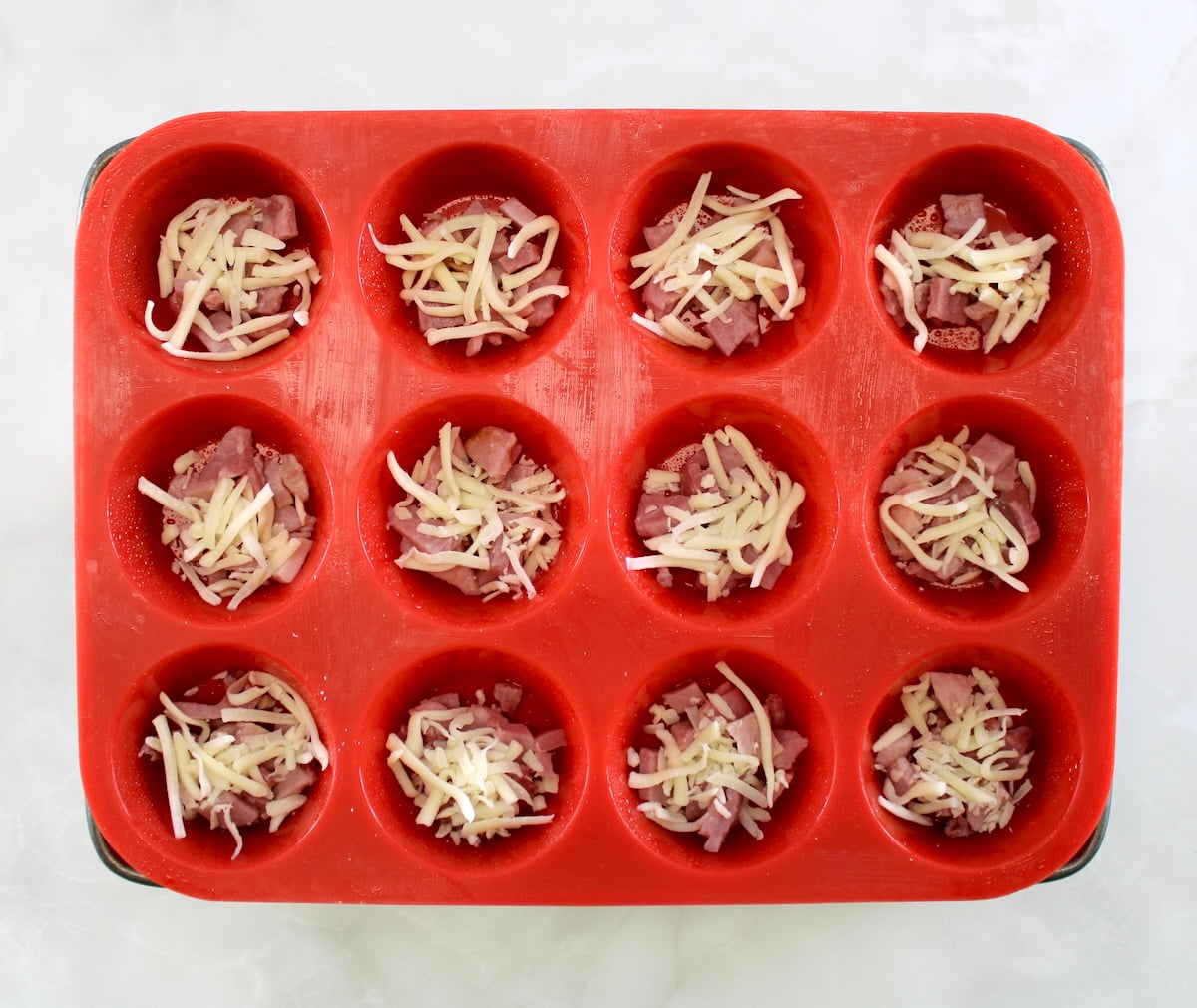 red silicone muffin pan with chopped ham and shredded cheese in each