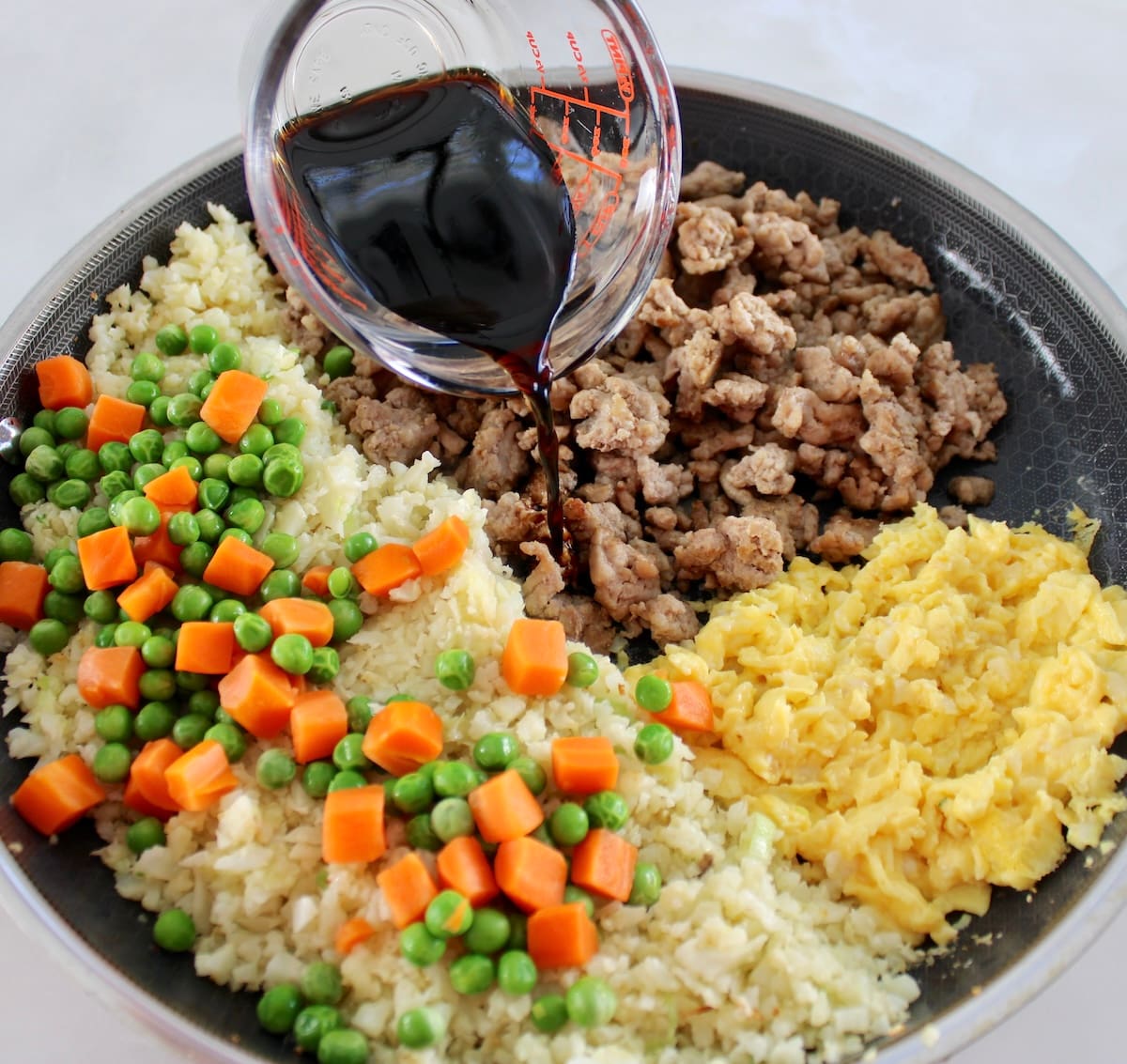 Pork Fried Cauliflower Rice ingredients in skillet in sections unmixed with liquid aminos pouring in