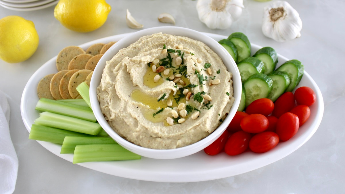 overhead closeup of Roasted Garlic Cauliflower Hummus in white bowl with fresh veggies and crackers around