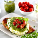 Grilled Chicken Margherita on white plate with pesto sauce and cherry tomatoes in back