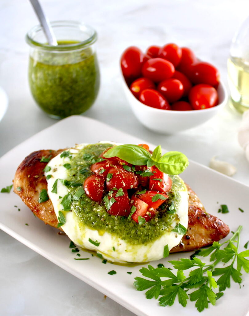 Grilled Chicken Margherita on white plate with pesto sauce and cherry tomatoes in back