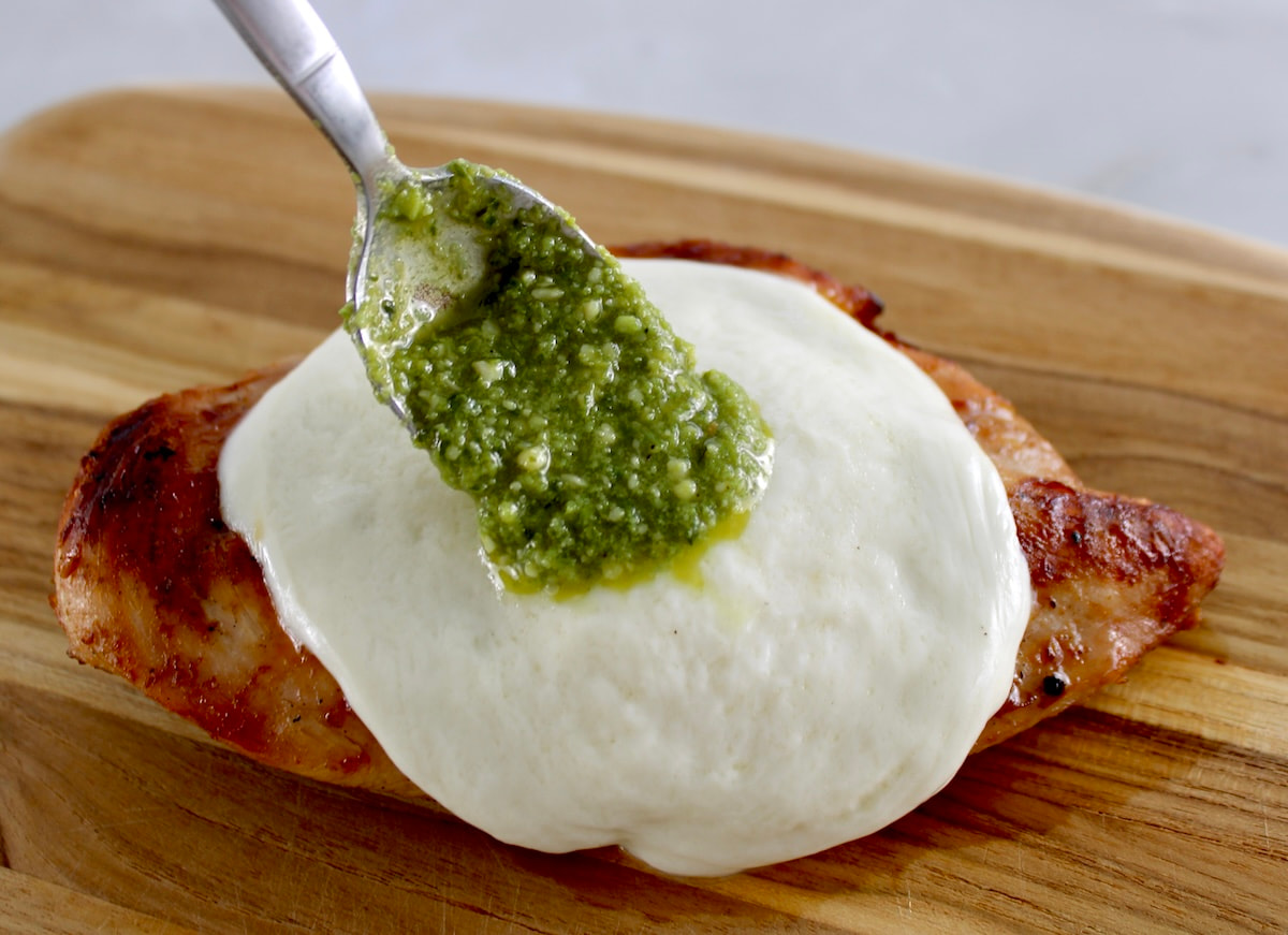 grilled piece of chicken on cutting board with melted mozzarella and pesto being spooned over top