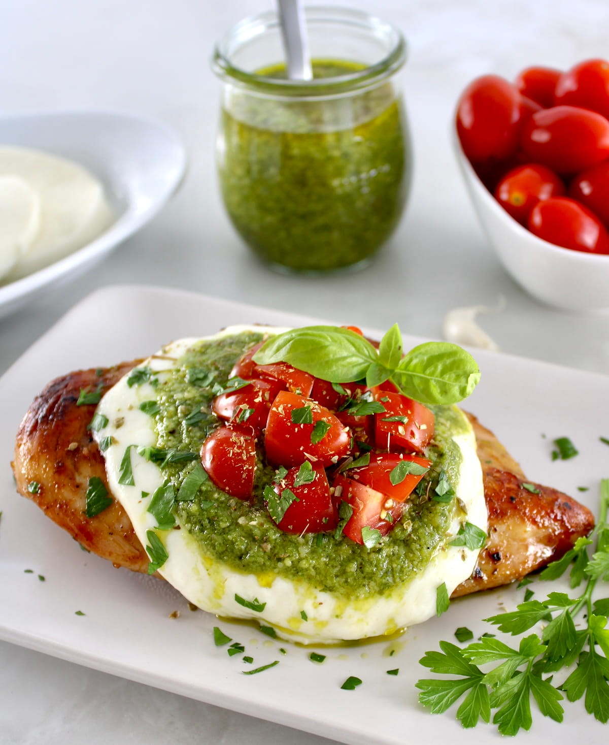 Grilled Chicken Margherita on white plate with pesto sauce and cherry tomatoes in back