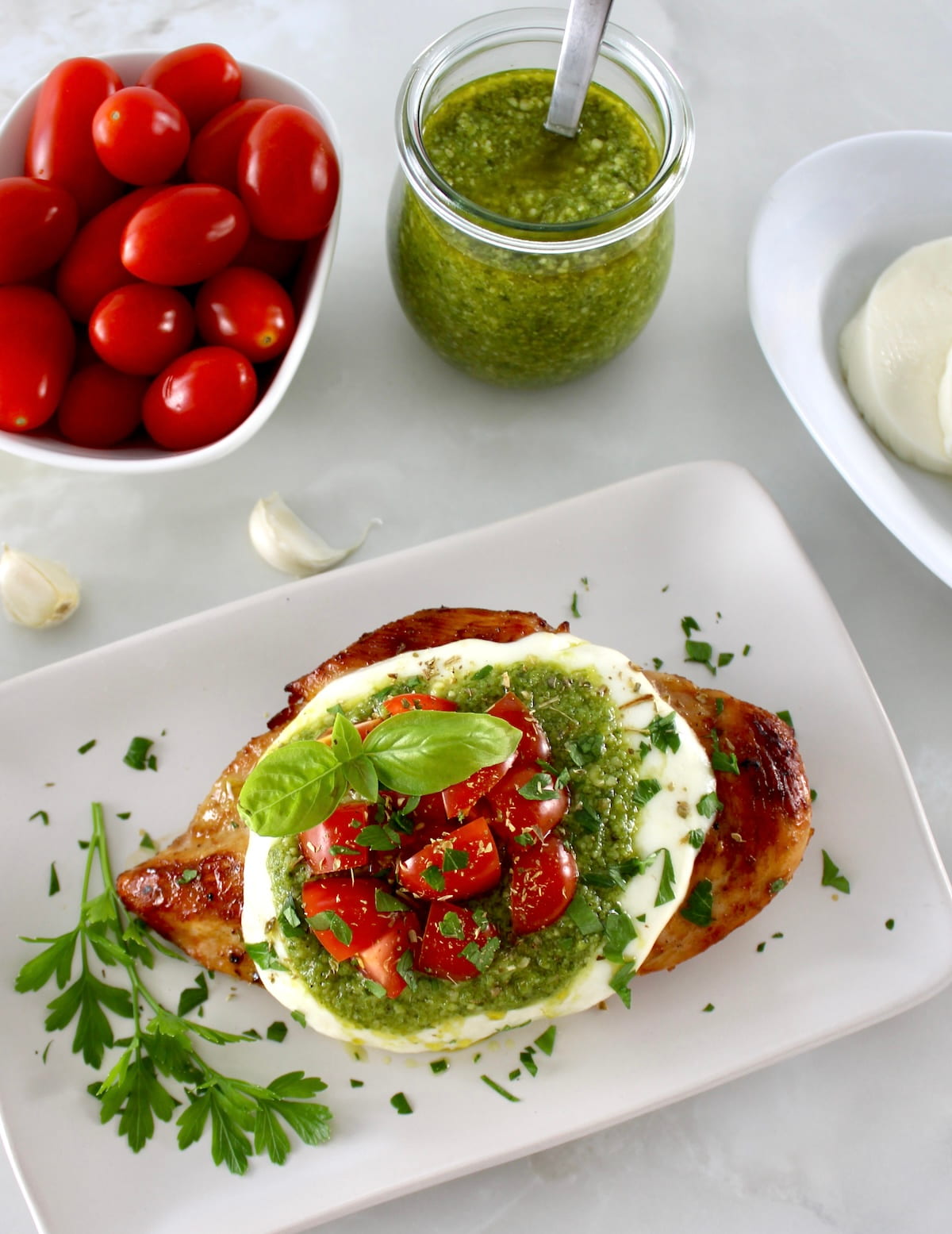 overhead view of Grilled Chicken Margherita on white plate with pesto and tomatoes in back