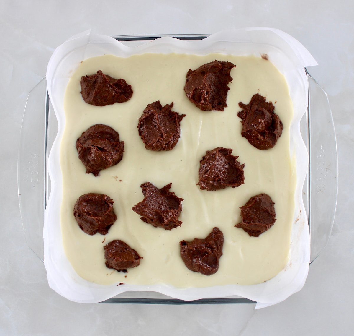 cheesecake batter in parchment lined glass baking dish with dollops of brownie batter on top