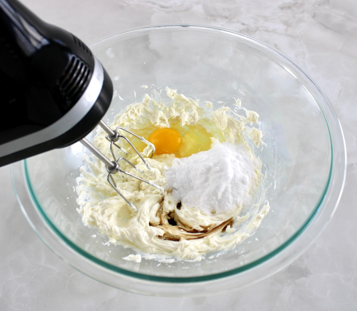 cream cheese, egg and sweetener in glass bowl being mixed with hand mixer