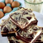 Keto Cheesecake Brownie Bars stacked up on white plate with eggs and almond flour and eggs in back