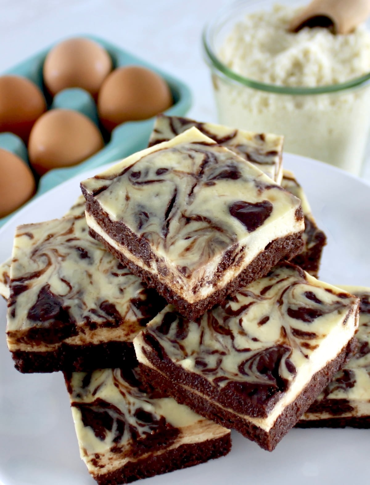Keto Cheesecake Brownie Bars stacked up on white plate with eggs and almond flour and eggs in back
