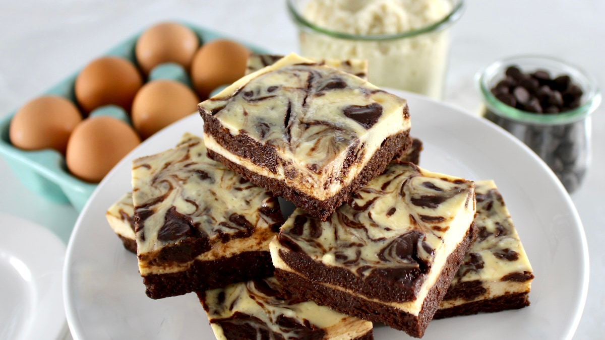 Keto Cheesecake Brownie Bars stacked up on white plate with eggs and almond flour in back