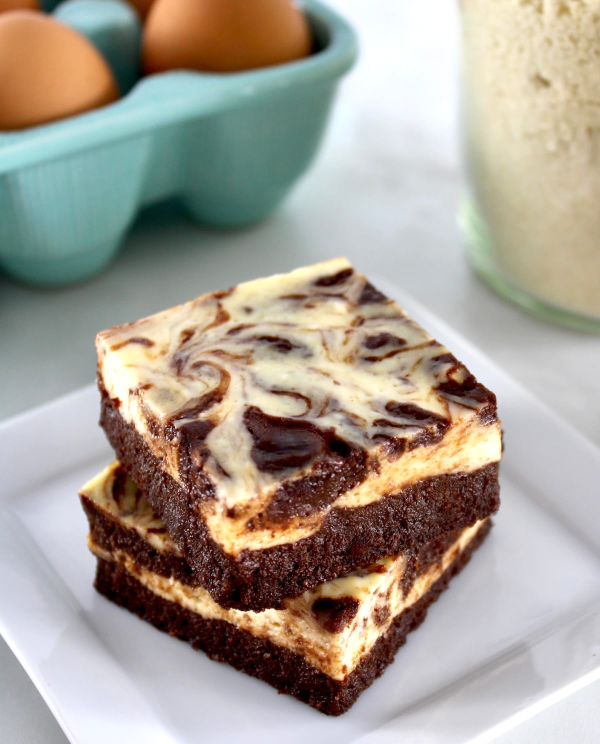 closeup of 2 Keto Cheesecake Brownie Bars stacked on white plate with eggs in back