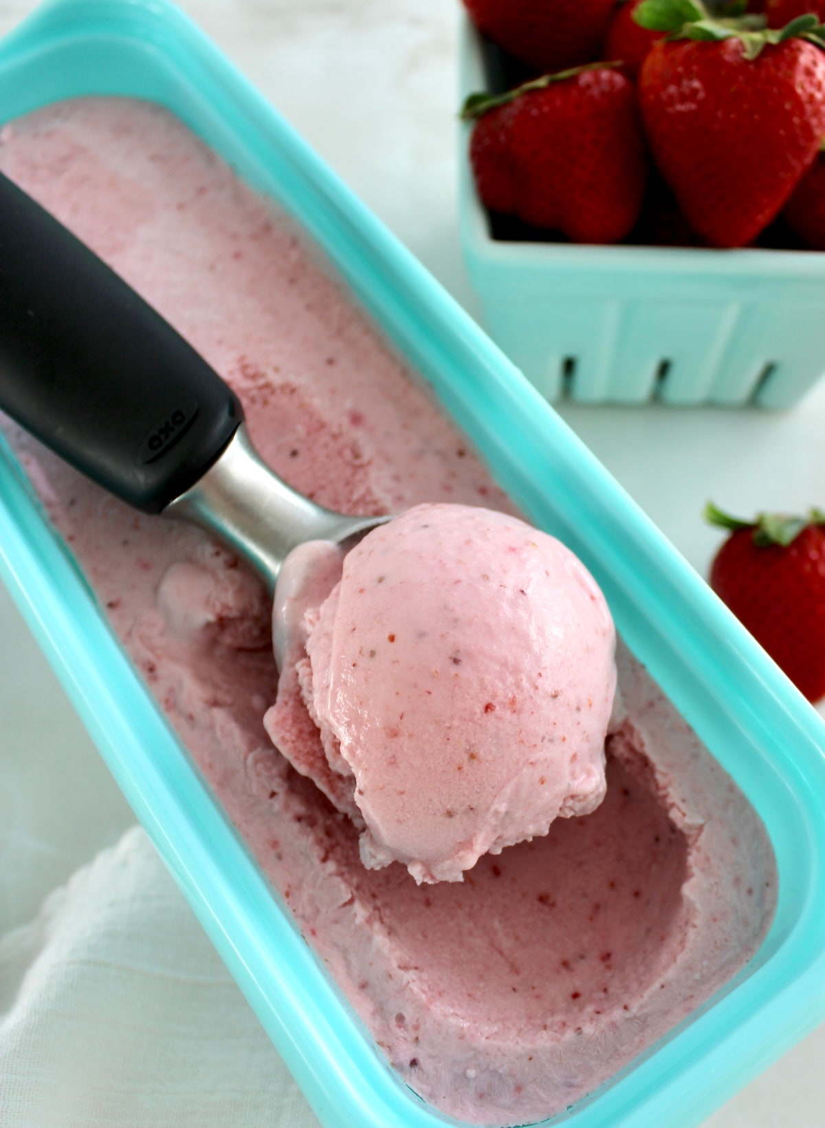 Strawberry Cottage Cheese Ice Cream in ice cream scoop resting in blue storage container with strawberries in back