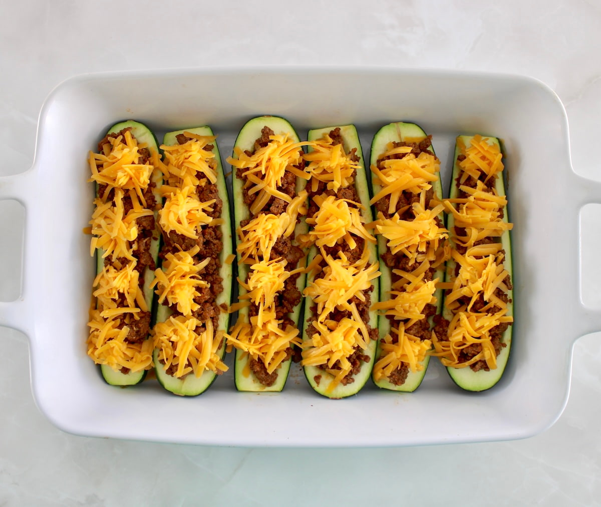 6 zucchini halves in white casserole filled with taco meat and shredded cheddar cheese on top
