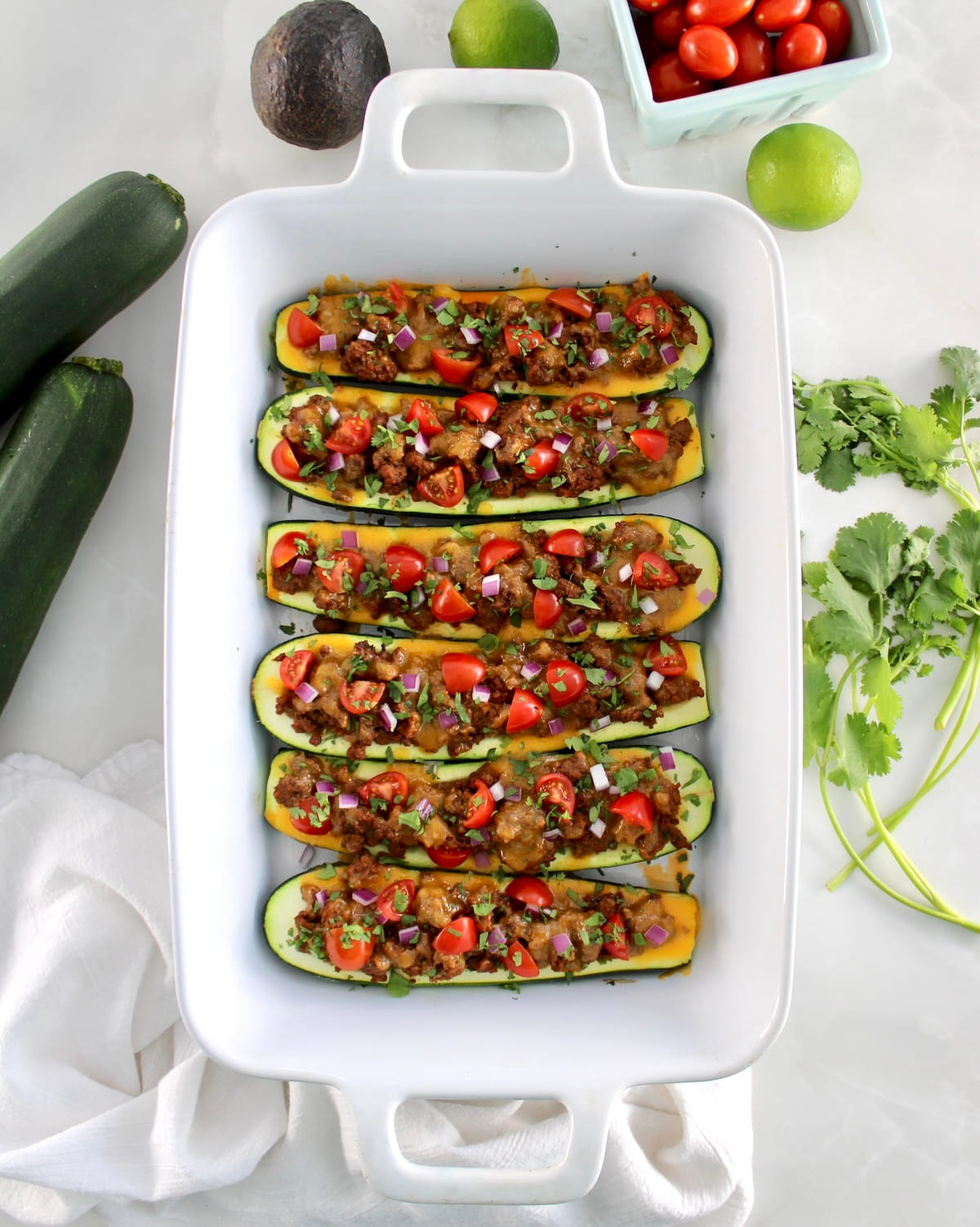 overhead view of 6 Taco Zucchini Boats in white casserole with fresh zucchini, tomatoes and cilantro around