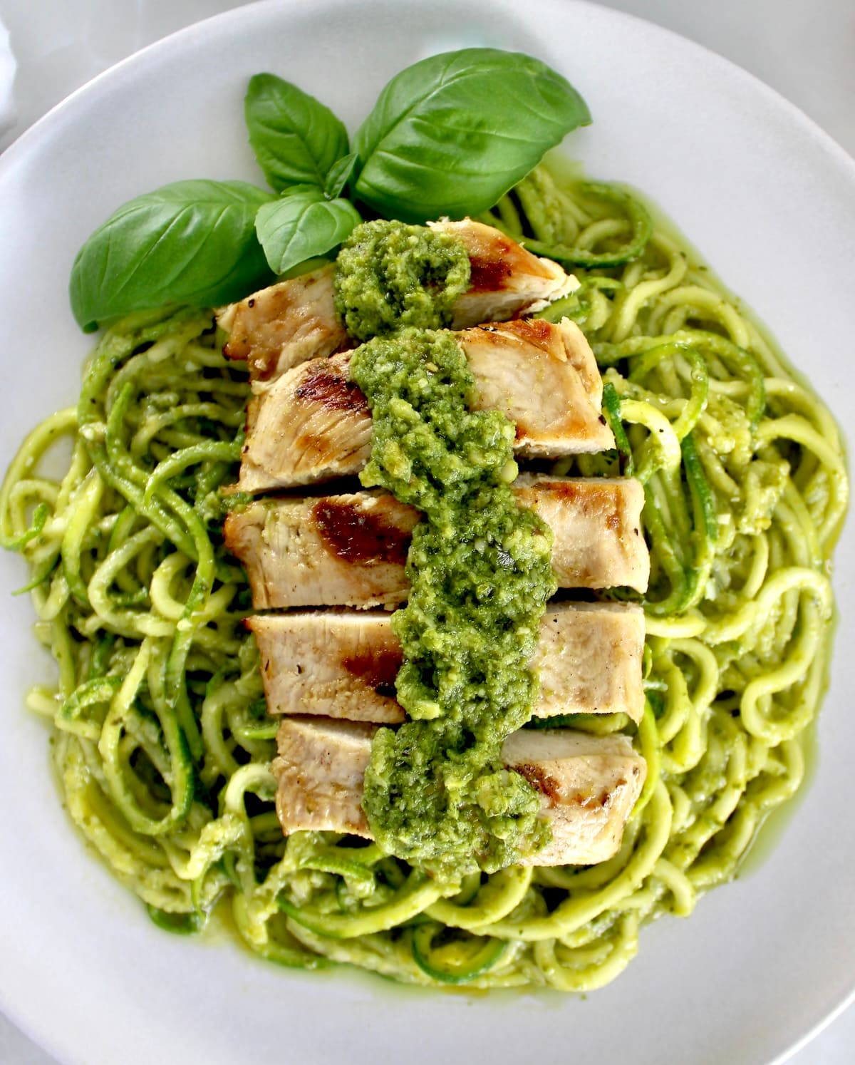 closeup of Grilled Lemon Chicken Pesto Zoodles on white plate with fresh basil garnish