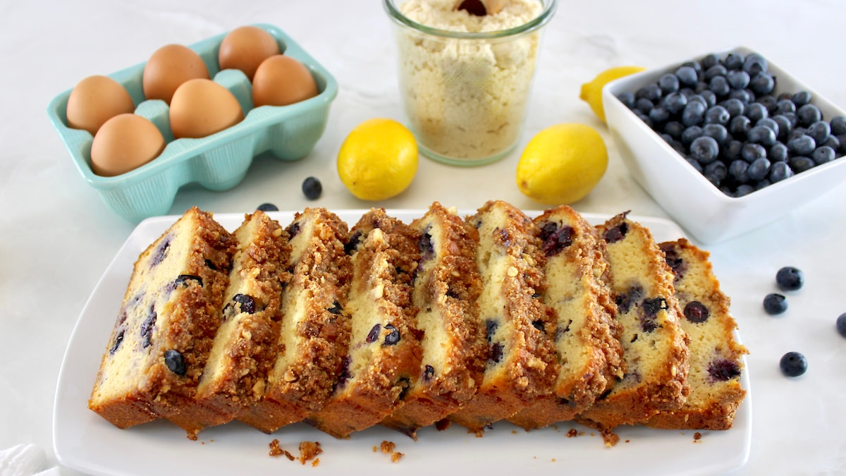 sliced Keto Blueberry Crumb Loaf on white platter with lemons, eggs and blueberries in back