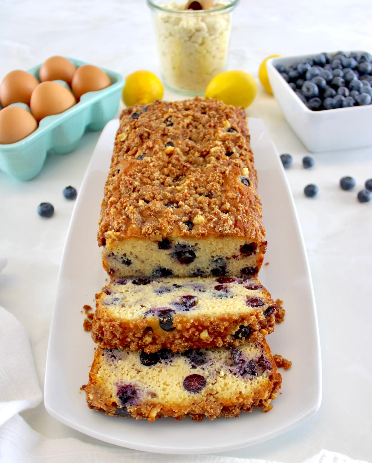 overhead view of Keto Blueberry Crumb Loaf on white platter and 2 slices cut
