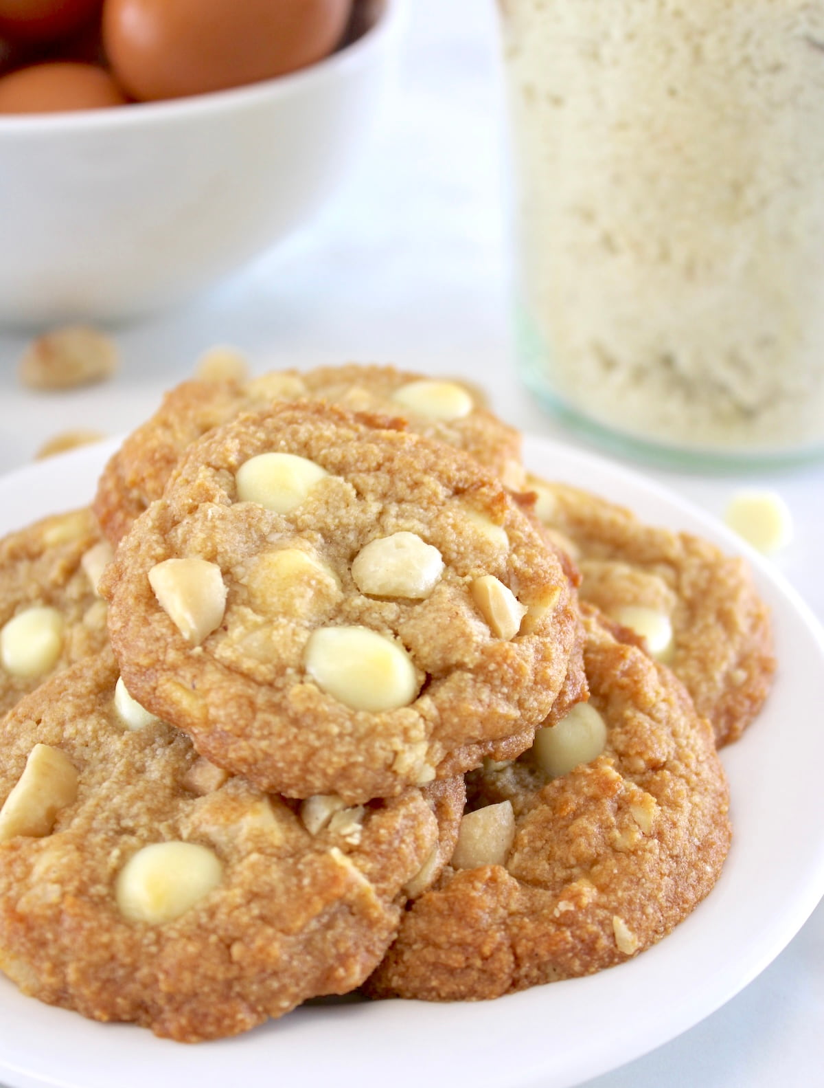closeup of Keto White Chocolate Macadamia Nut Cookies on white plate with almond flour and eggs in back