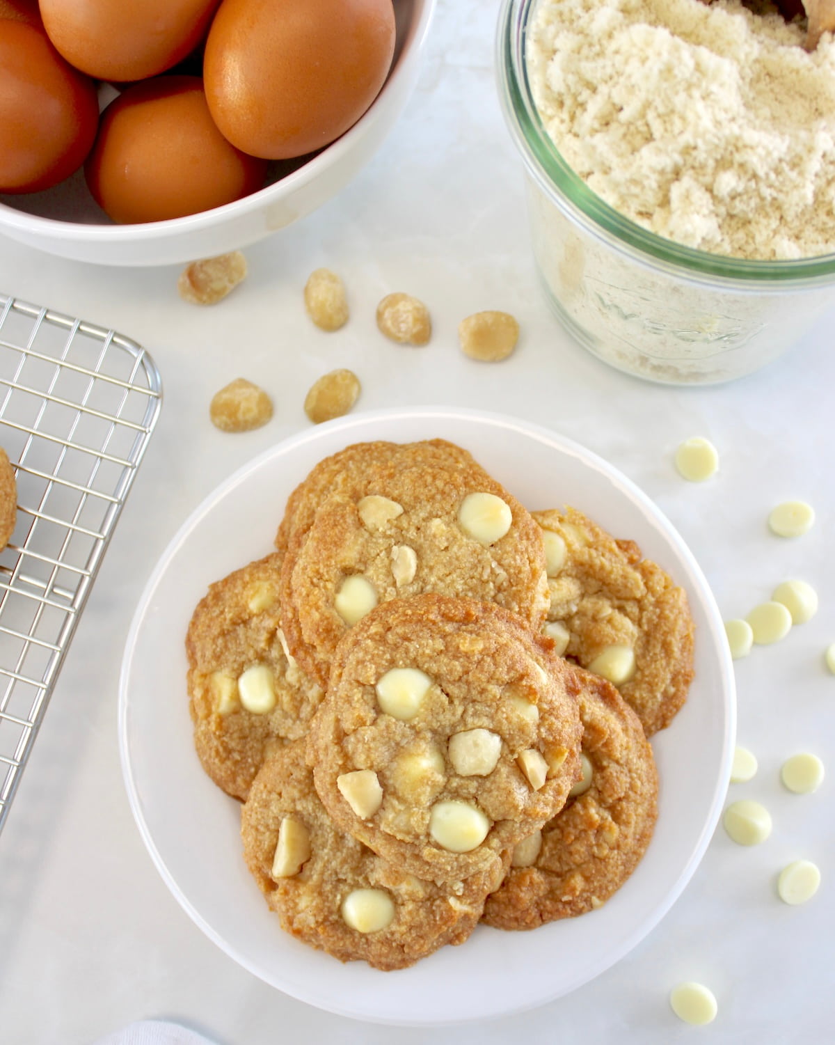 overhead view of Keto White Chocolate Macadamia Nut Cookies on white plate with almond flour and eggs in back