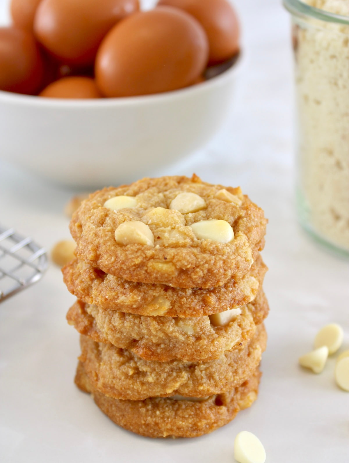  Keto White Chocolate Macadamia Nut Cookies stacked up with eggs and almond flour in back