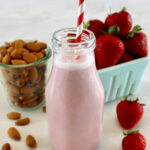 Strawberry Almond Milk in glass bottle with red and white straw, strawberries and almonds in back