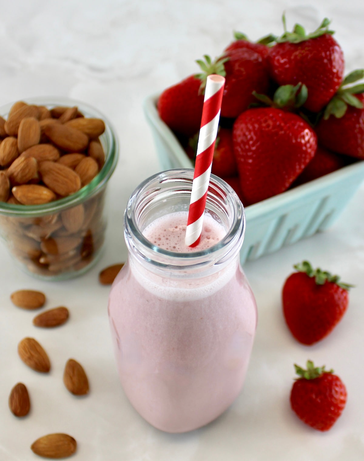 Strawberry Almond Milk in glass bottle with red and white straw, strawberries and almonds in back