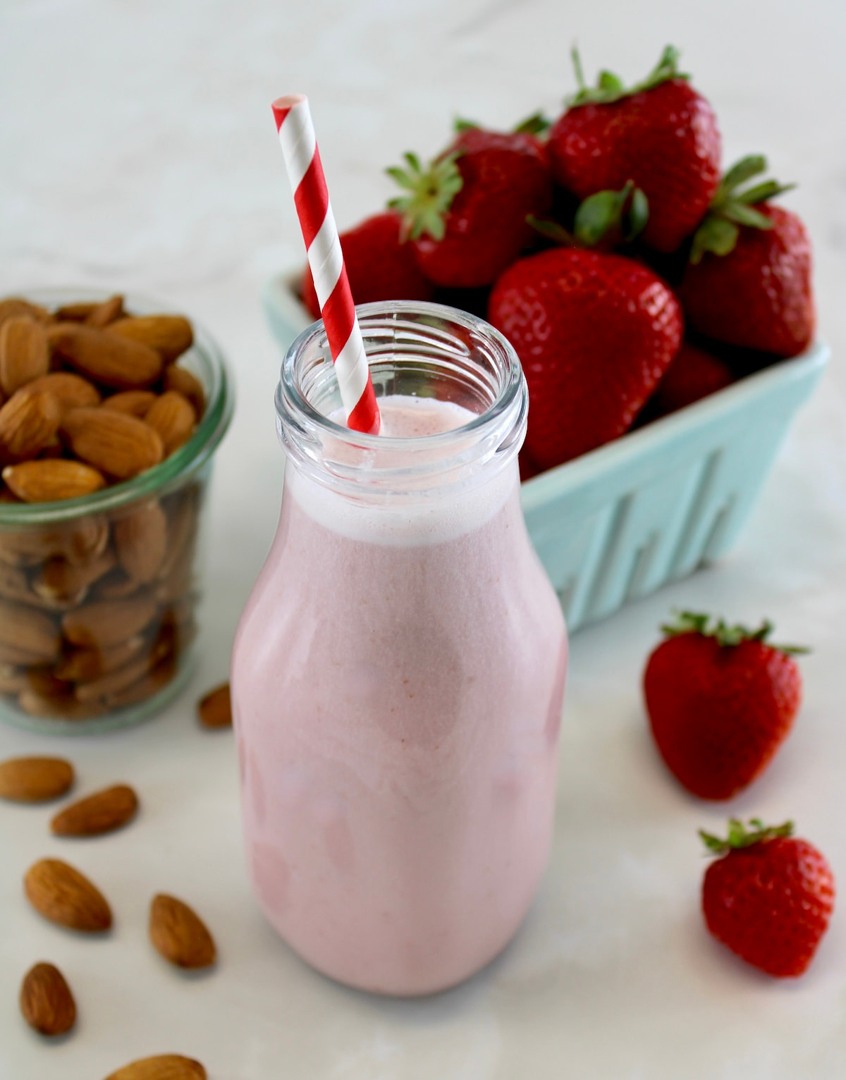 Strawberry Almond Milk in glass bottle with red and white straw, strawberries and almonds in back