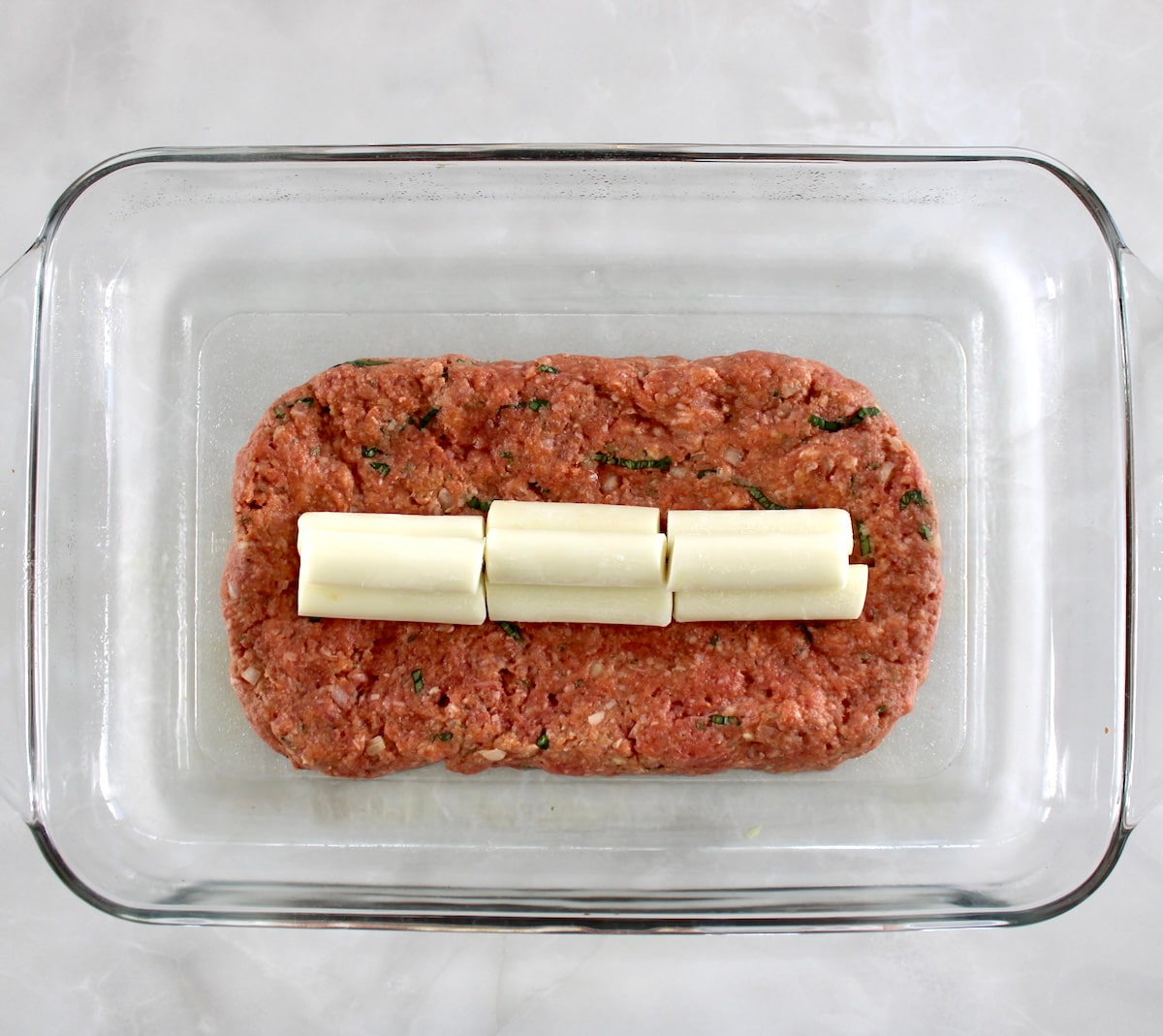 Bacon Wrapped Cheese Stuffed Italian Meatloaf mixture with pieces of frozen string cheese on top in middle