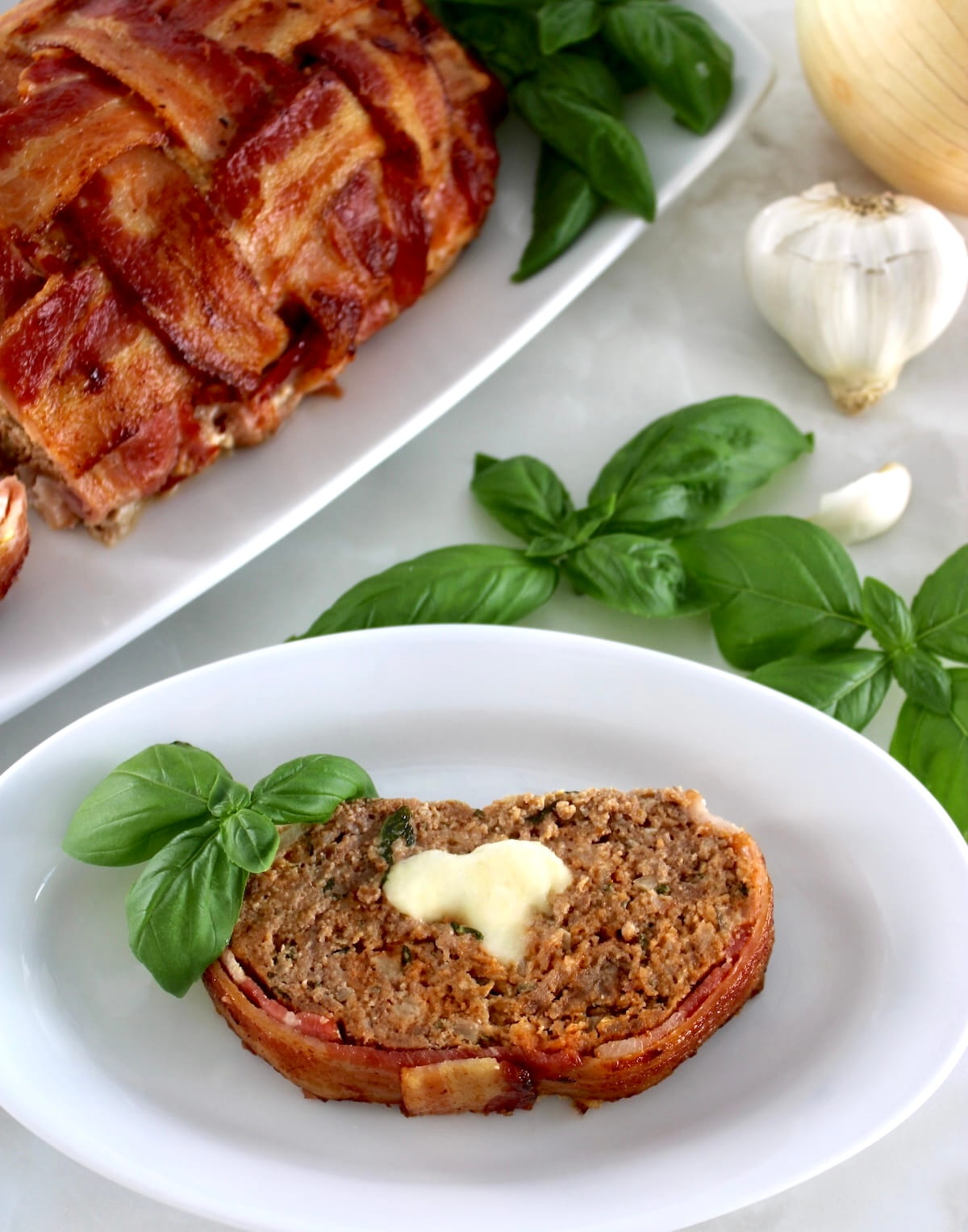 overhead view of slice of Bacon Wrapped Cheese Stuffed Italian Meatloaf on white plate with fresh basil
