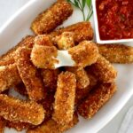keto mozzarella cheese sticks on white platter with marinara on side and one cheese stick pulled apart
