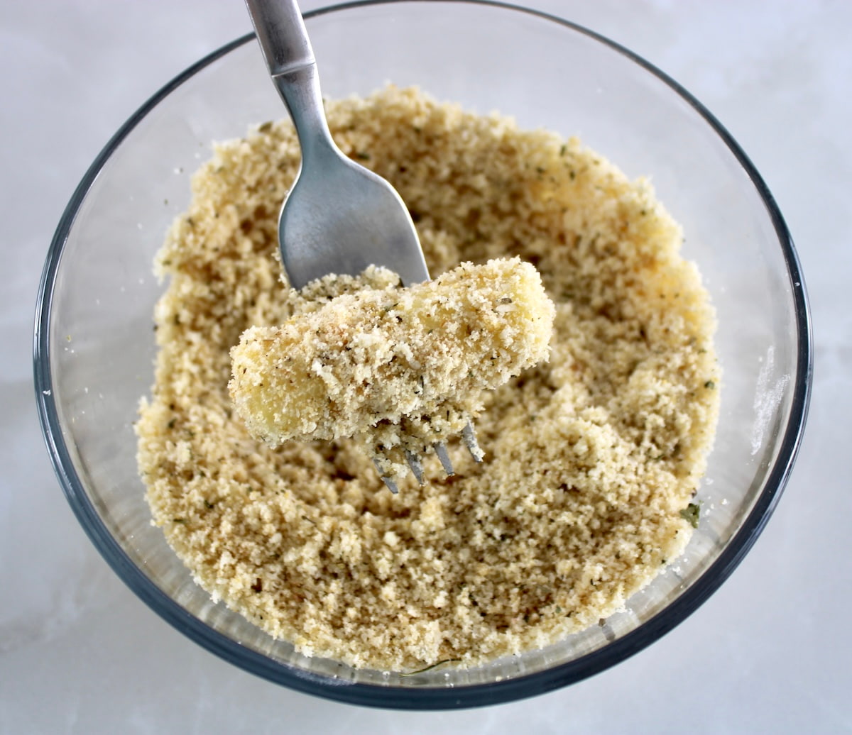mozzarella cheese stick being dipped into breading with fork in glass bowl