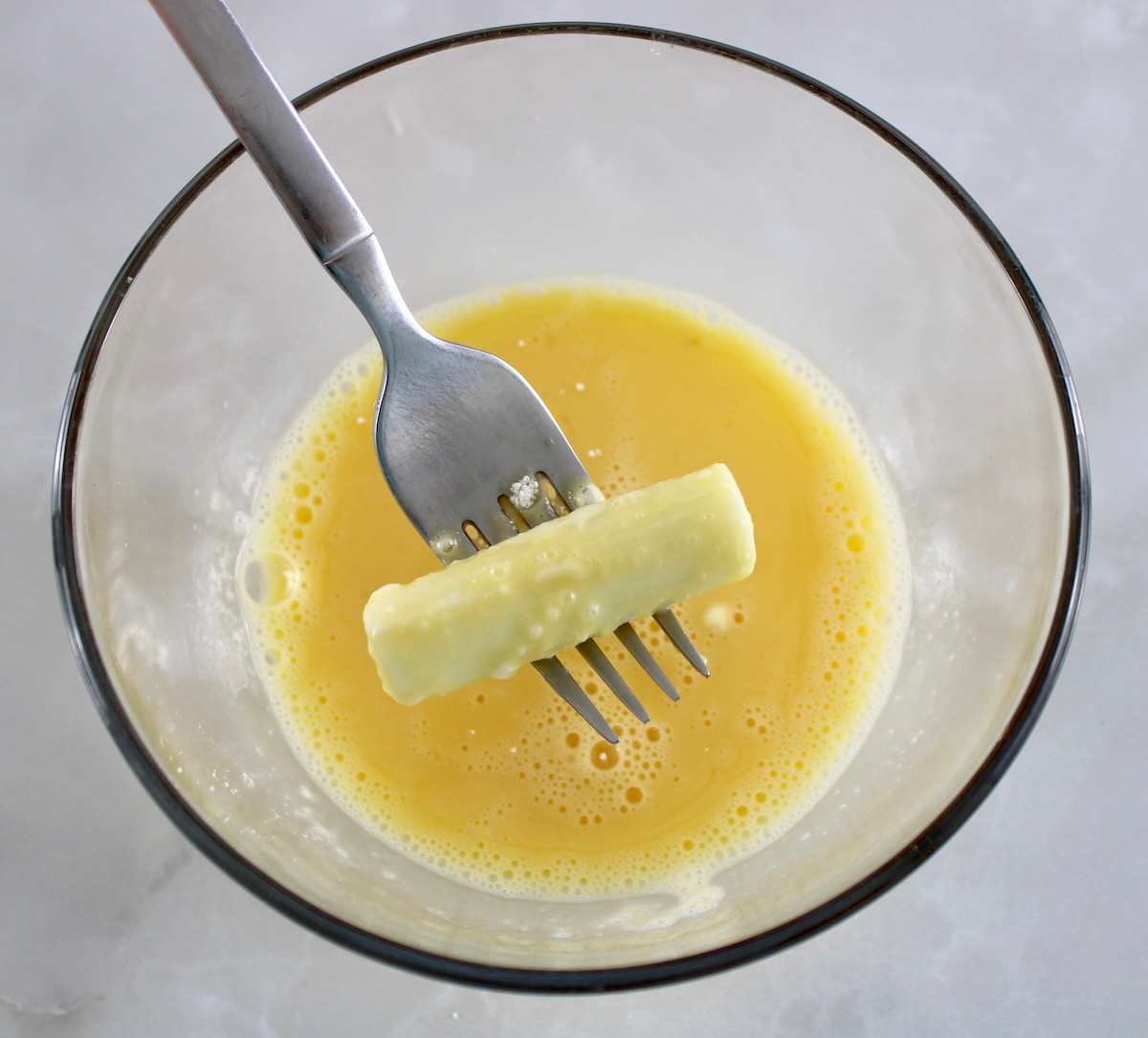 mozzarella cheese stick being dipping into beaten eggs with fork in glass bowl