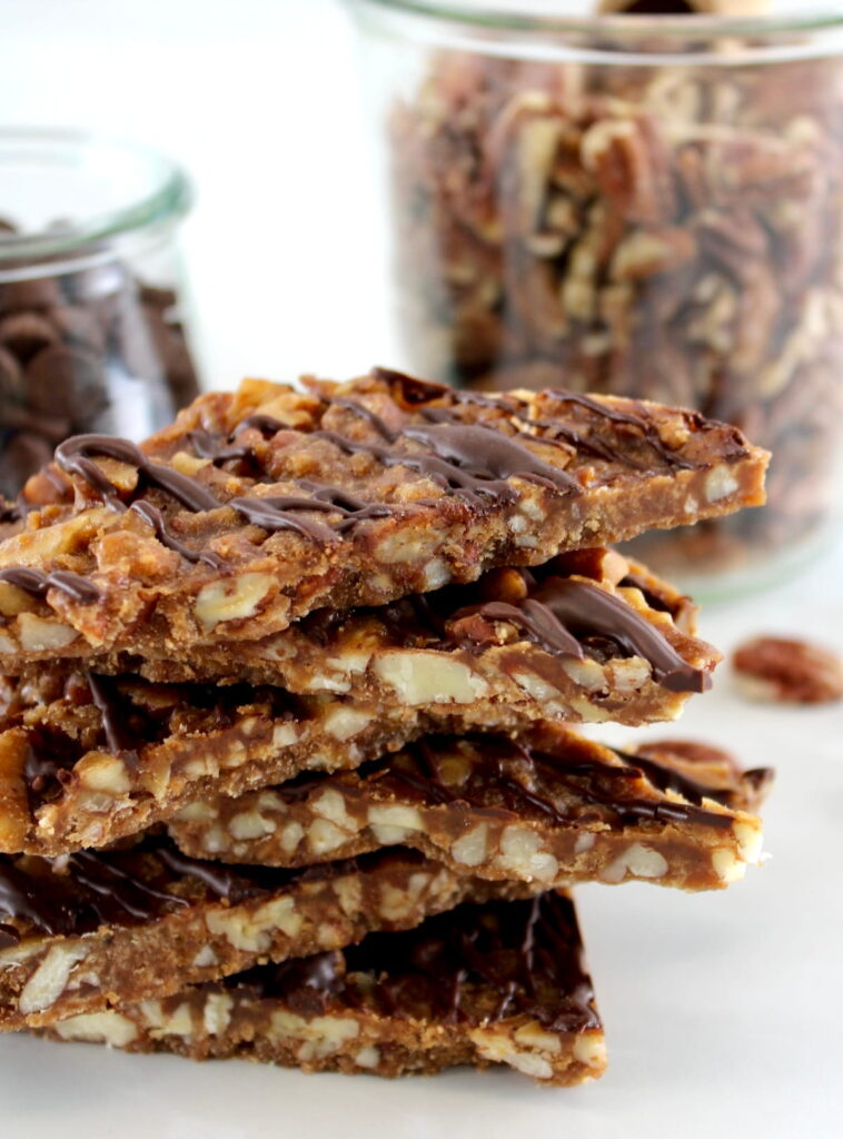 Keto Pecan Praline Bark pieces stacked up with pecans and chocolate chips in jars in back