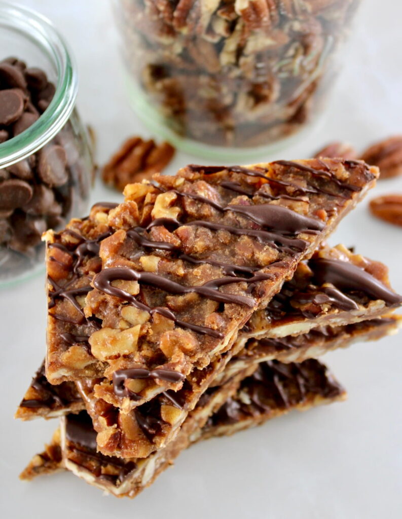 closeup of Keto Pecan Praline Bark stacked up with chocolate chips and pecans in jars behind