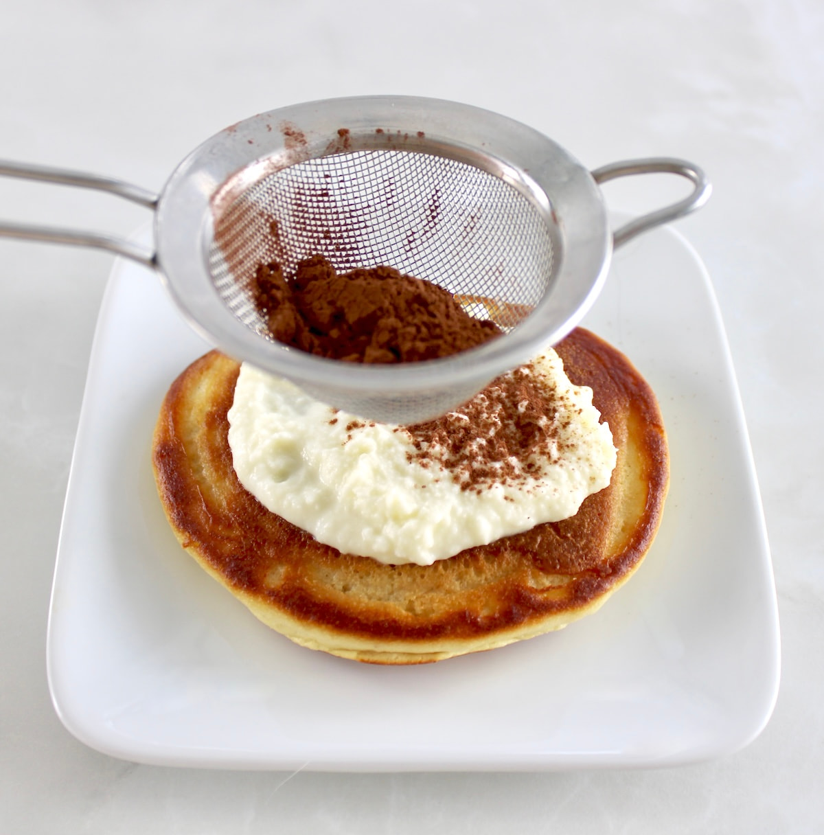 pancake on white plate with mascarpone cream on top and cocoa powder being sifted on top