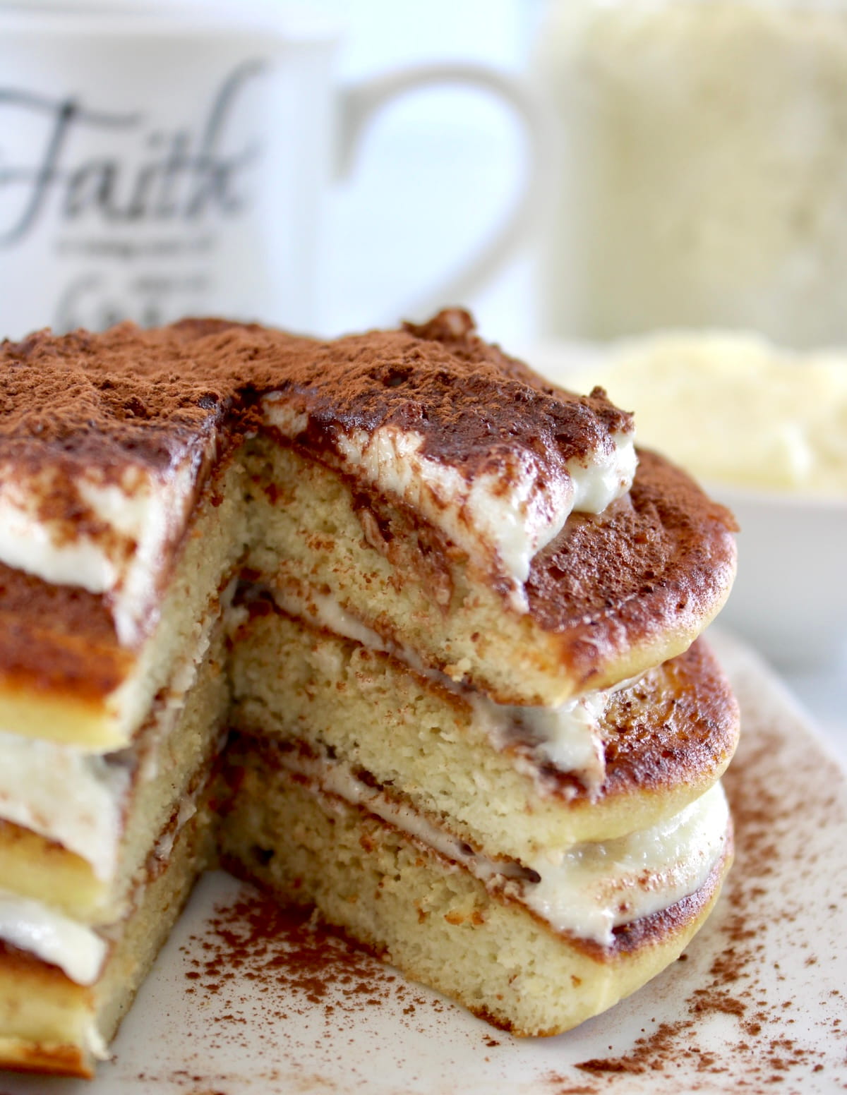 closeup of Keto Tiramisu Pancakes stacked on white plate with mug of coffee in back with piece missing