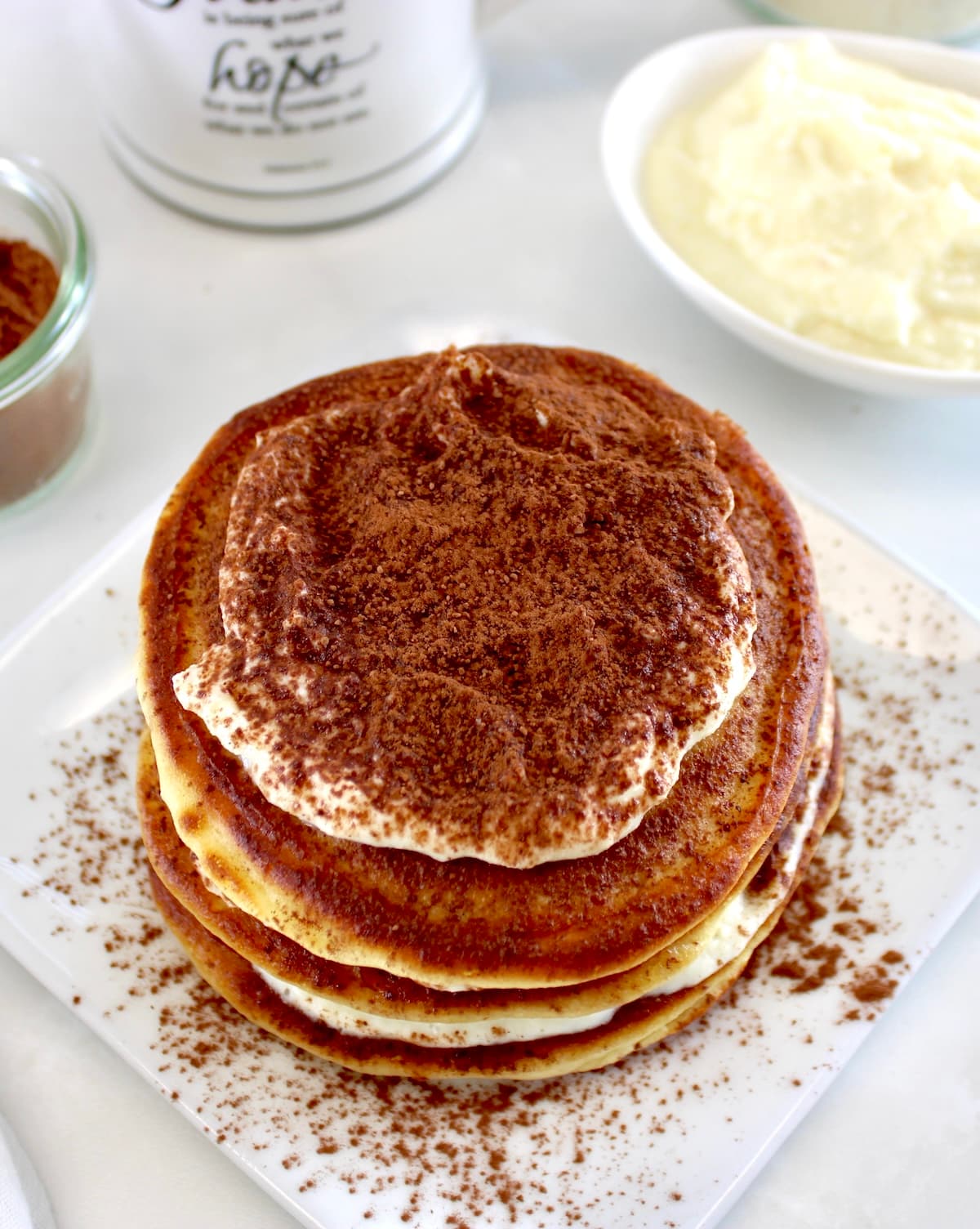 overhead view of Keto Tiramisu Pancakes stacked on white plate with mug of coffee in back on white plate