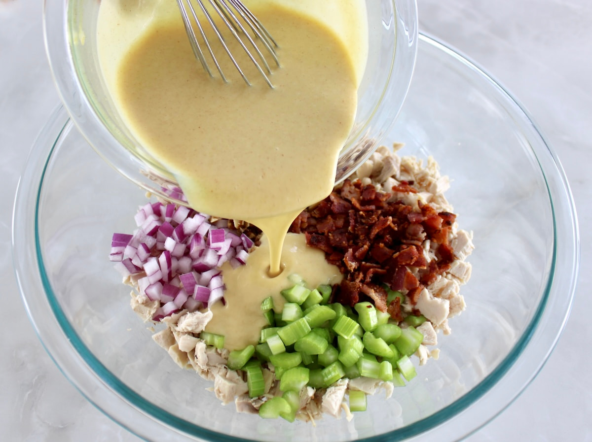 honey mustard dressing being poured over chicken salad ingredients in glass bowl unmixed