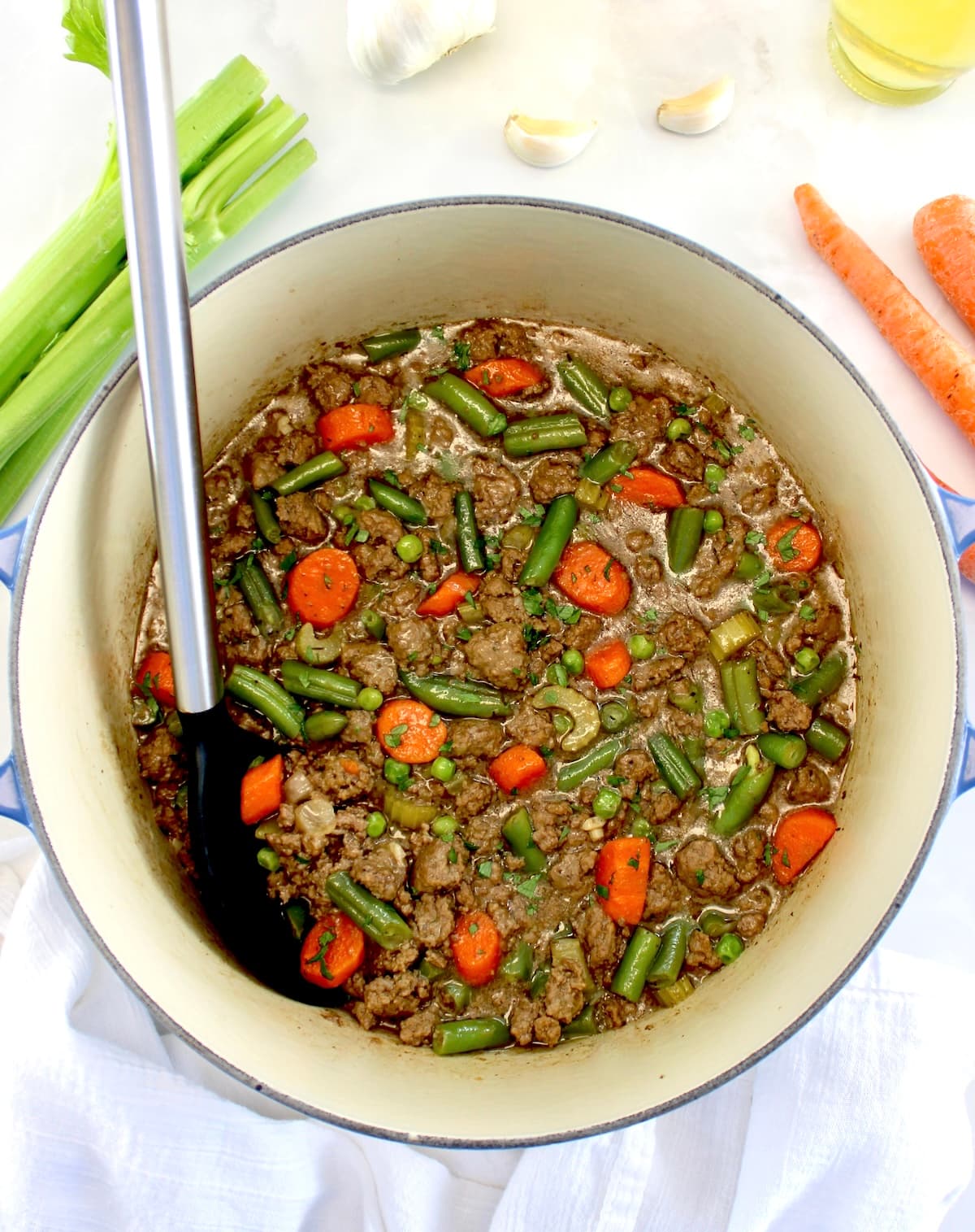 overhead view of Ground Beef Stew in pot with black serving spoon on side and veggies in background