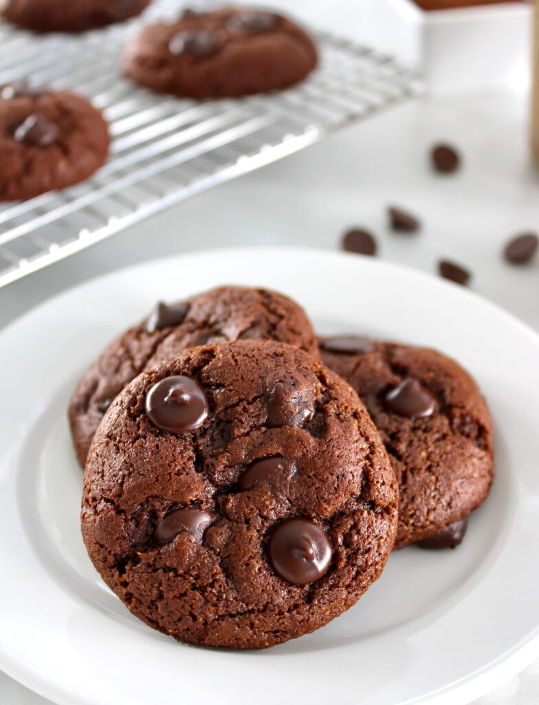 3 Keto Chocolate Almond Butter Cookies on white plate with more in back on cooling rack