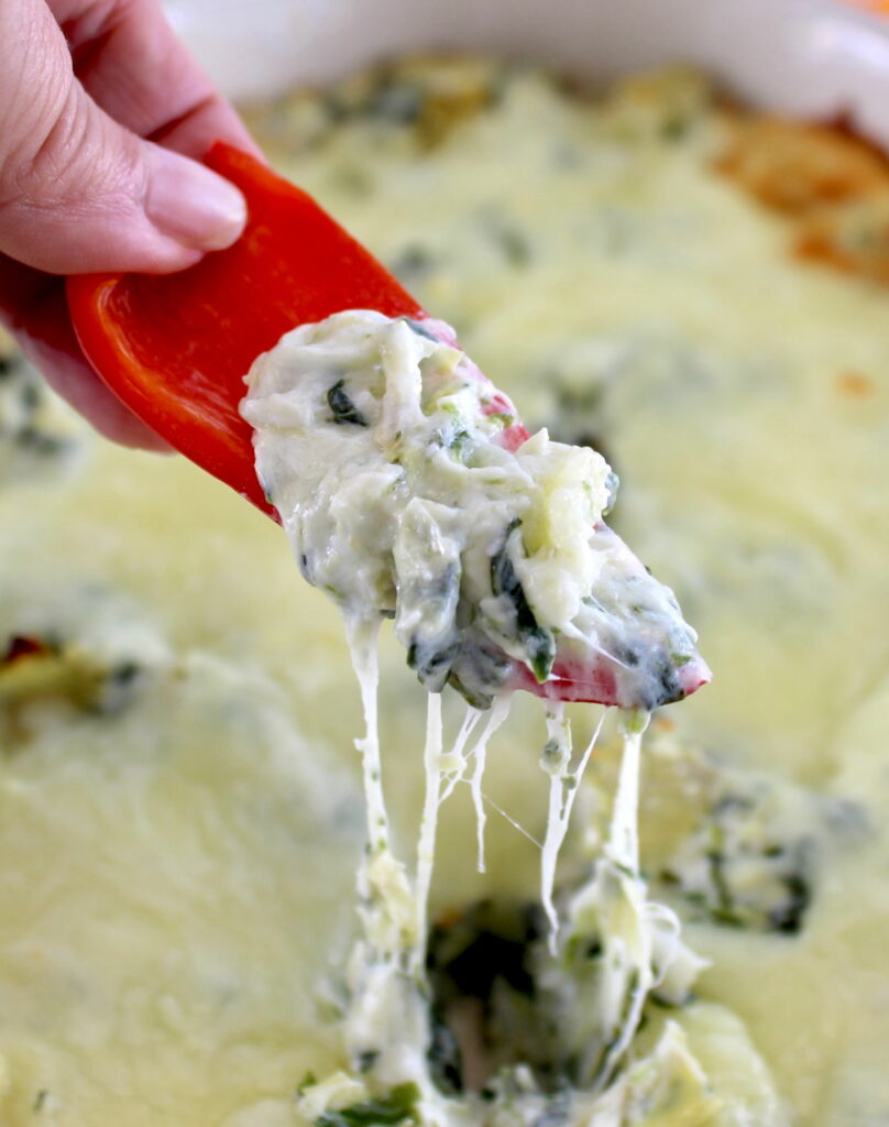 holding red bell pepper dipped in Spinach Artichoke Dip