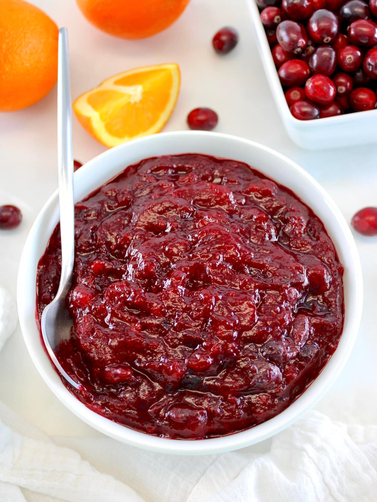 Keto Cranberry Sauce in white bowl with spoon on side with cranberries and oranges in back