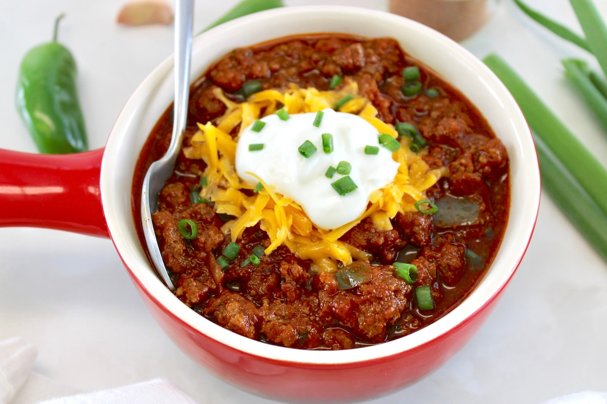 Keto Instant Pot No-Bean Chili in red crock with spoon on side, shredded cheddar and sour cream on top